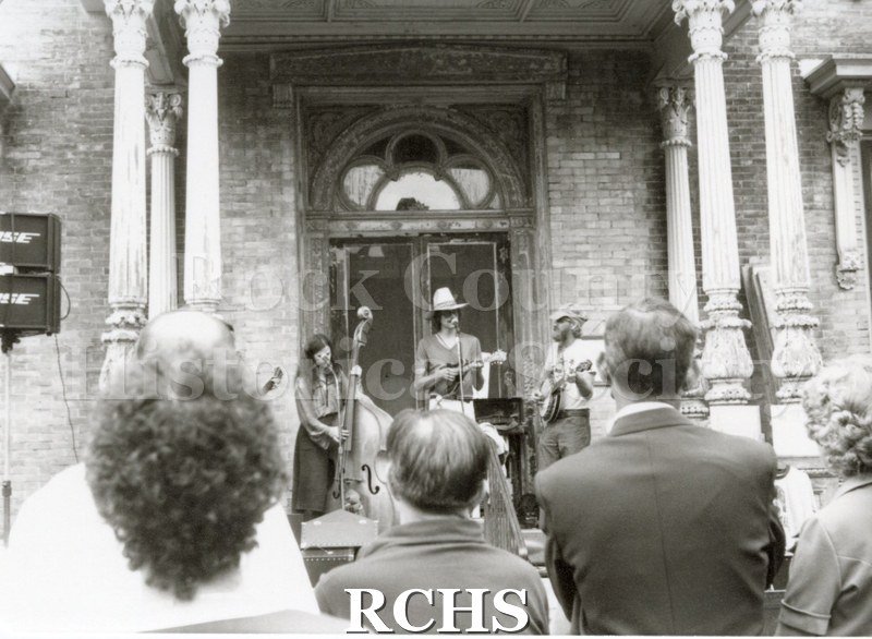 Black and white photograph of the Tallman Arts Exhibit. People are listening to a band playing on the porch of the Tallman House.
