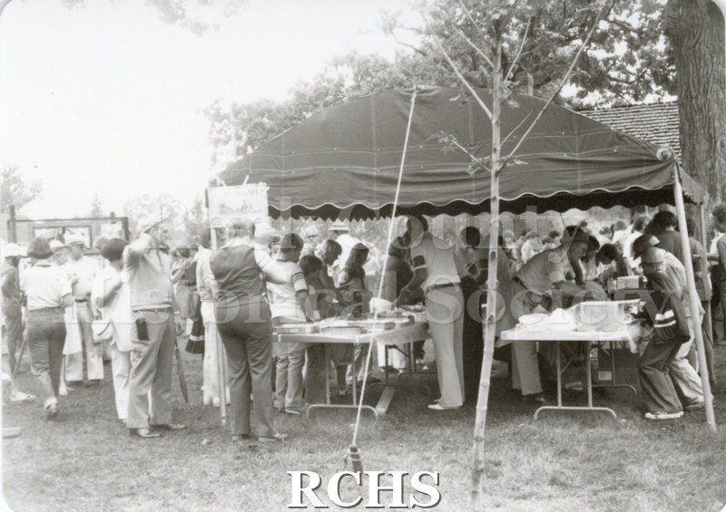Black and white photograph of the Tallman Arts Exhibit. People are standing around the tables to buy ice cream and cake.
