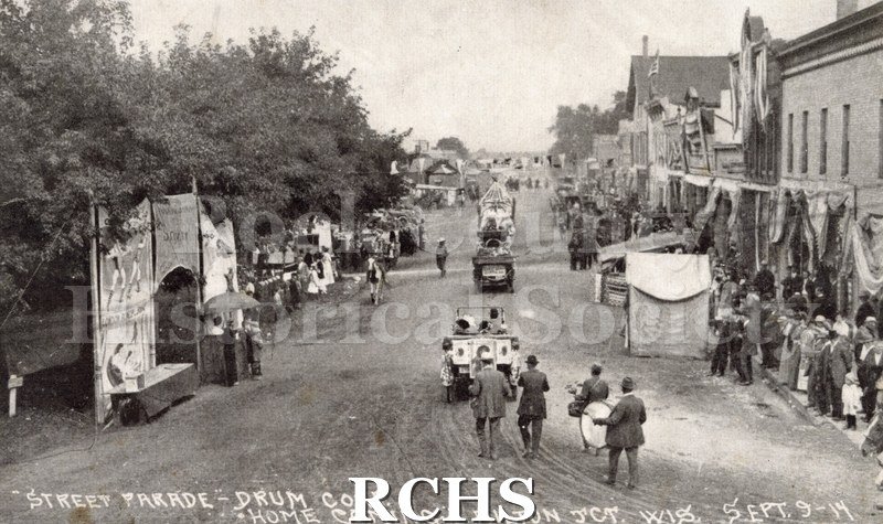 Black and white photograph of a parade in Milton Junction. There are people standing on the sidewalk viewing the parade. On bottom: "Street Parade-Drum Corp.