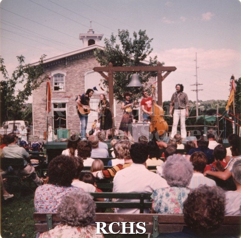 Color photo of the Stage One Band performing in front of the Coach House with an audience seated in front ot them.