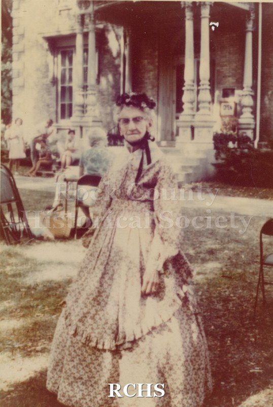 Tinted photo of Mrs. Inez Smith at the Tallman House. Shows an older woman standing in front of the Tallman House. She is dressed in a period costume.