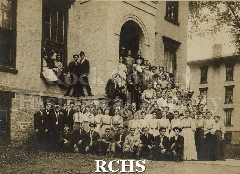 Black and white photograph of several young people sitting and standing in front of a building.