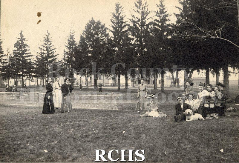 Black and white photograph of Milton College students. Several students are sitting and standing near a tennis court.