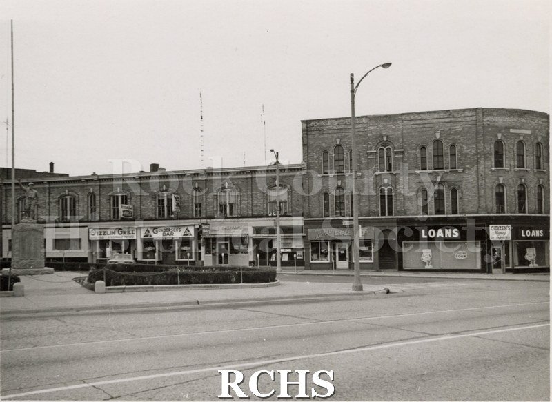 Black and white photograph of Corn Exchange in Janesville. Shows businesses on the block: such as, Loans; Gunderson's Bar; Shephard's Jewelry and Sizzlin Grilll.