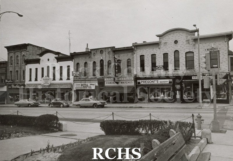 Black and white photograph of stores on Milwaukee street. They include: Pregont's Bakery; Smith Jewelry; Villa Pizza; and Yagla's.