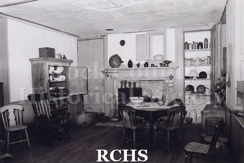 Black and white photograph of the Tallman House basement kitchen. Shows the table with a bowl on it and chairs around it; the fireplace and hutch.