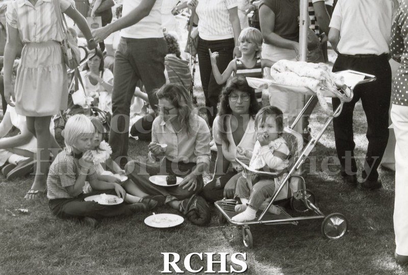 Black and white photograph of the 1983 Tallman Festival. Shows families sitting on the ground eating.