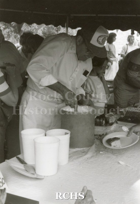 Black and white photograph of the 1983 Arts Festival showing volunteers serving food . One man is scooping ice cream out of a container.