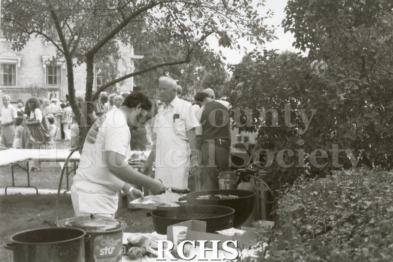 Black and white photograph of volunteers serving food at the Arts Festival. One man is working on a grill.