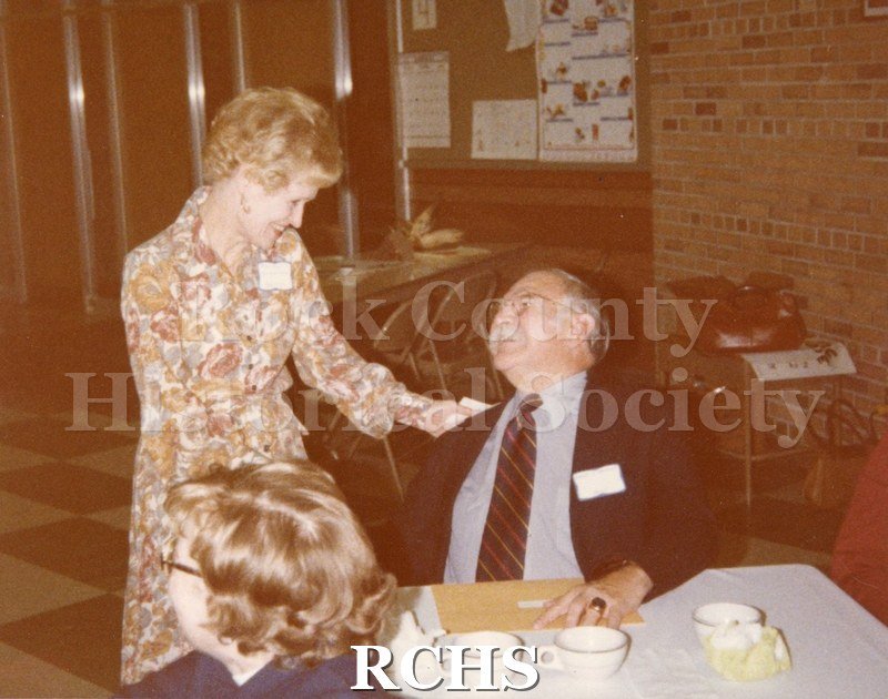 Color photo of Dorothy Gilbertson as she leans down to speak to Dr.Robert Inmann who is seated at a table.