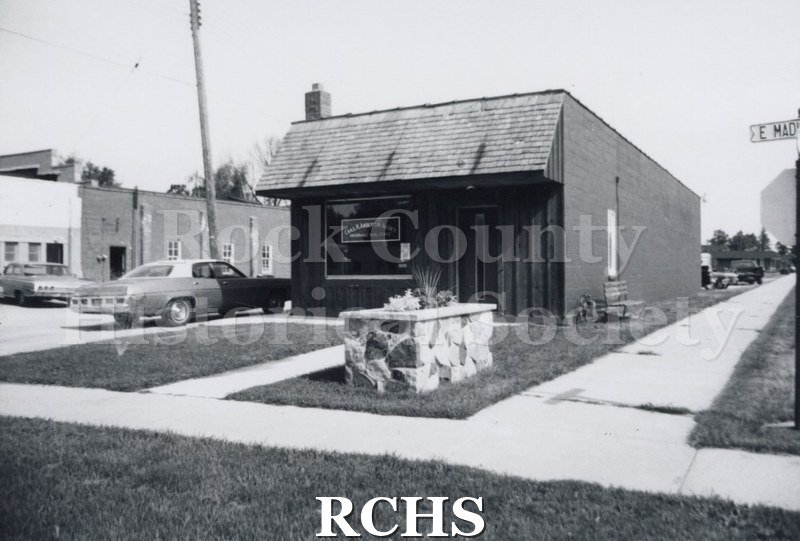 Black and white photograph of an unidentified business in Milton. There is a car parked on the left side and a planter in the grass in front of the building.