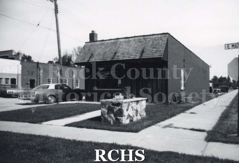 Black and white photograph of an unidentified business in Milton. There is a car parked on the left side and a planter in the grass in front of the building.
