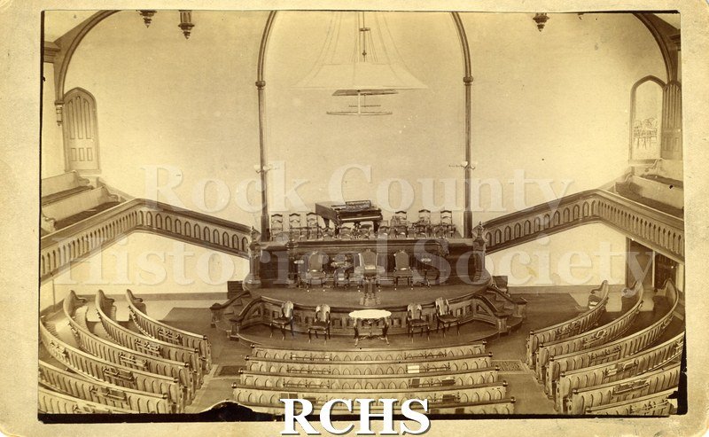 Black and white photograph of the Congregational Church, Janesville, WI. Shows the inside of a church with a grand piano on stage with chairs around it.
