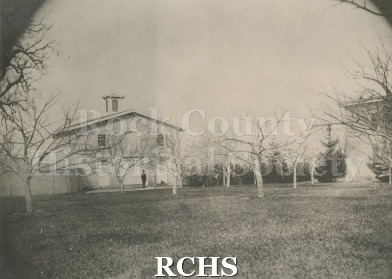 Black and white photograph of an 1870 view of Carriage House. Shows fence, orchard and W. M. Tallman.