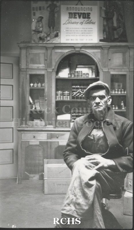 Black and white photograph of Alphonse Spangler sitting on a chair in front of a display cabinet.