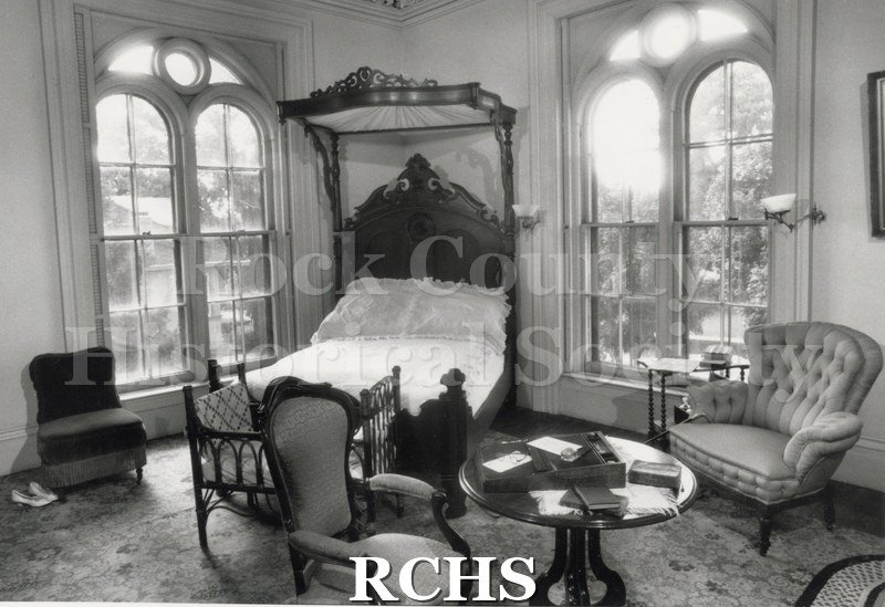 Black and white photograph of a bedroom in the Tallman House. Has a bed in the corner with a chair beside the window on the right side and a table in the front.