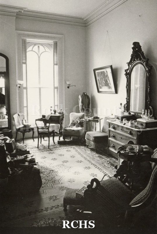 Black and white photograph of a bedroom in the Tallman House. Shows the three drawer dresser with pier mirror; a chair in the corner and a table in front of the.