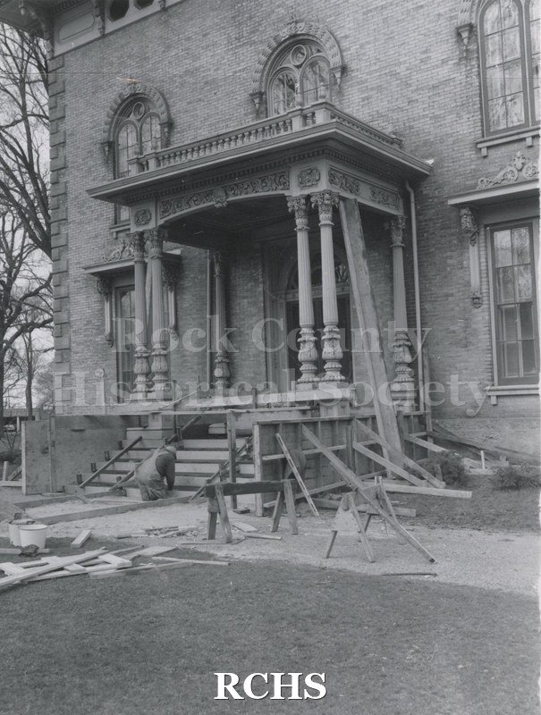 Black and white photograph of the front of the Tallman House. There is a workman on his knees working on the steps and boards are visible on the driveway and on the.
