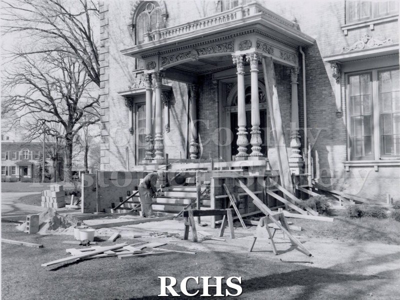 Black and white photograph of the front of the Tallman House. There is a workman on his knees working on the steps and boards are visible on the driveway and on the.
