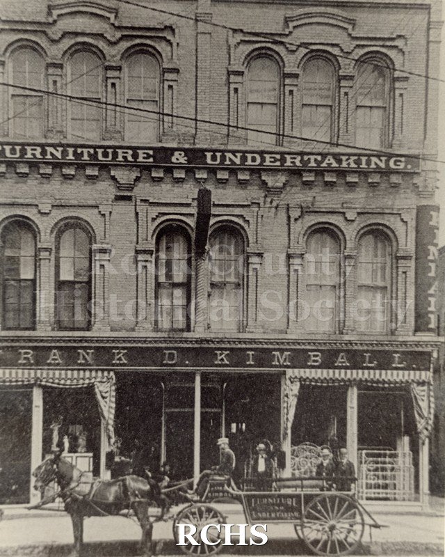 Black and white photograph of a three story building with "Frank D. Kimball" on the bottom and "Furniture & Undertaking" on the second floor.