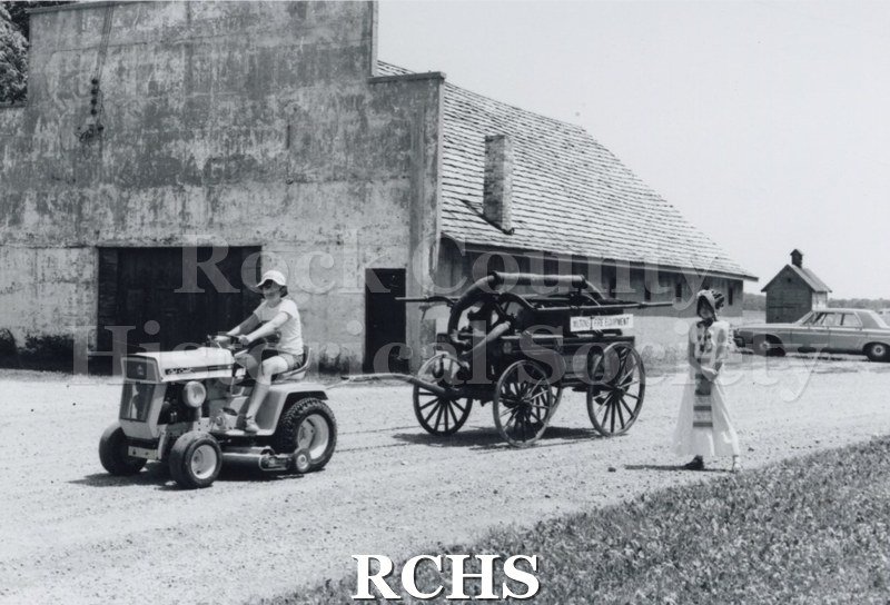 Black and white photograph of Glenn & Melanie Watson. The man is riding a tractor pulling an old fire wagon. A woman in period dress is walking beside him.
