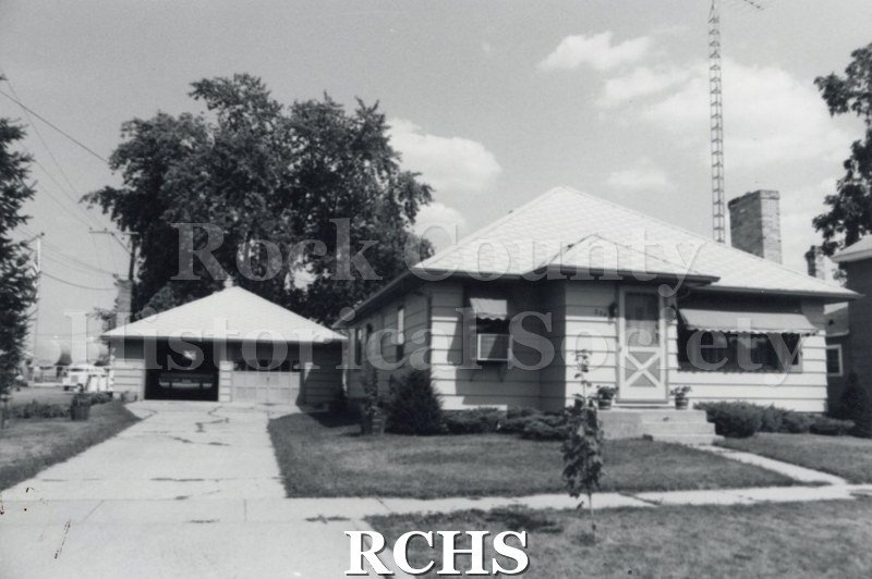 Black and white photograph of an unidentified one story house with awnings over the windows and a two door detached garage.