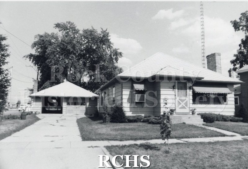 Black and white photograph of an unidentified one story house with awnings over the windows and a two door detached garage.