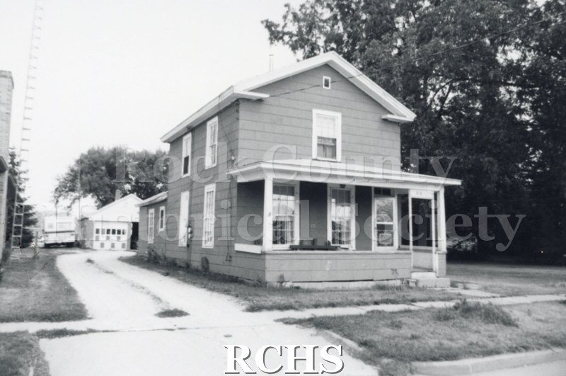 Black and white photograph of an unidentified two story house with a front porch and a detached garage in back.