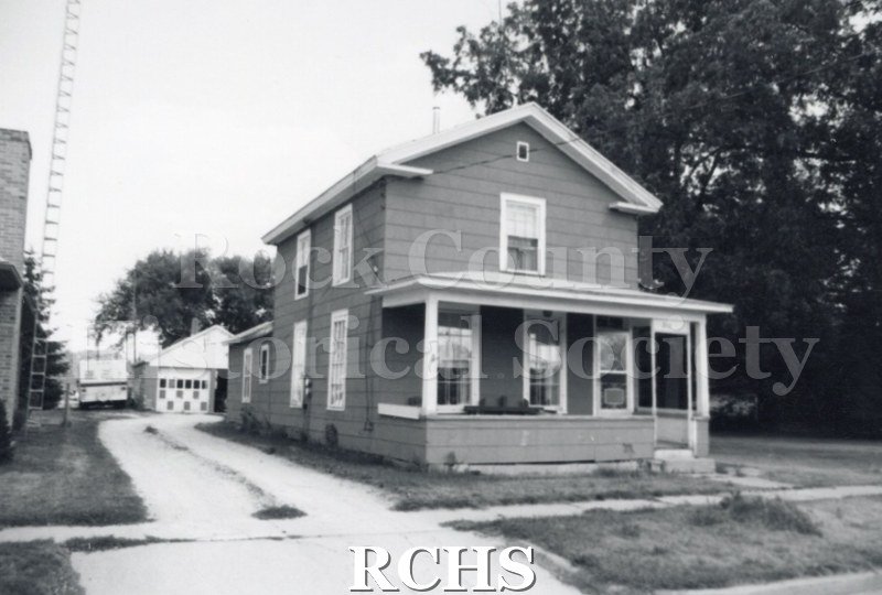 Black and white photograph of an unidentified two story house with a front porch and a detached garage in back.