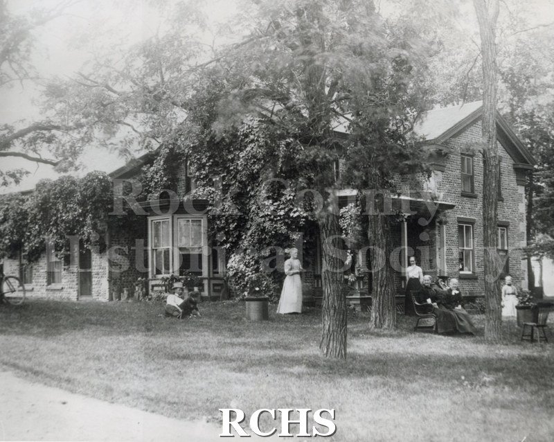 Black and white photograph of the Samuel Jones Cobblestone House, Clinton, WI, a two story brick house with people sitting and standing around it.