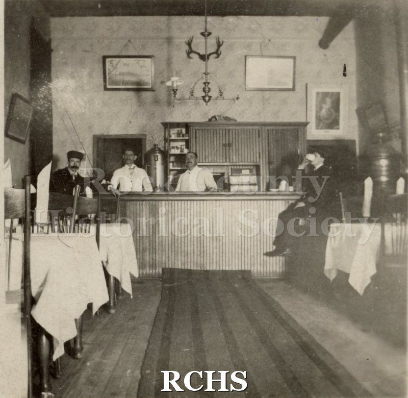 Black and white photograph of the inside of Myers Hotel cafe. There is a man drinking coffee on the right; two men behind the counter and a man standing on the left.