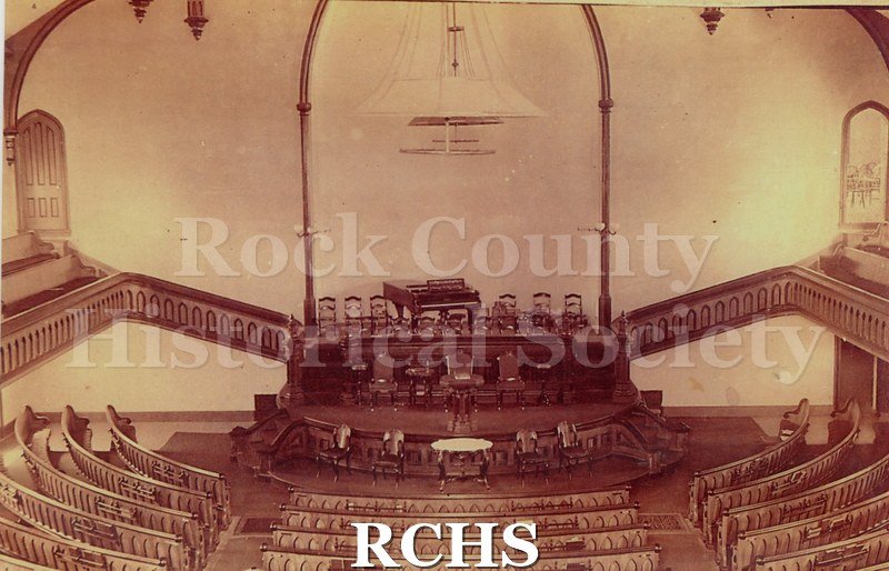 Color photo of the interior of an unidentified church. There are pews in a half circle in the front and a grand piano on a platform with chairs around it.