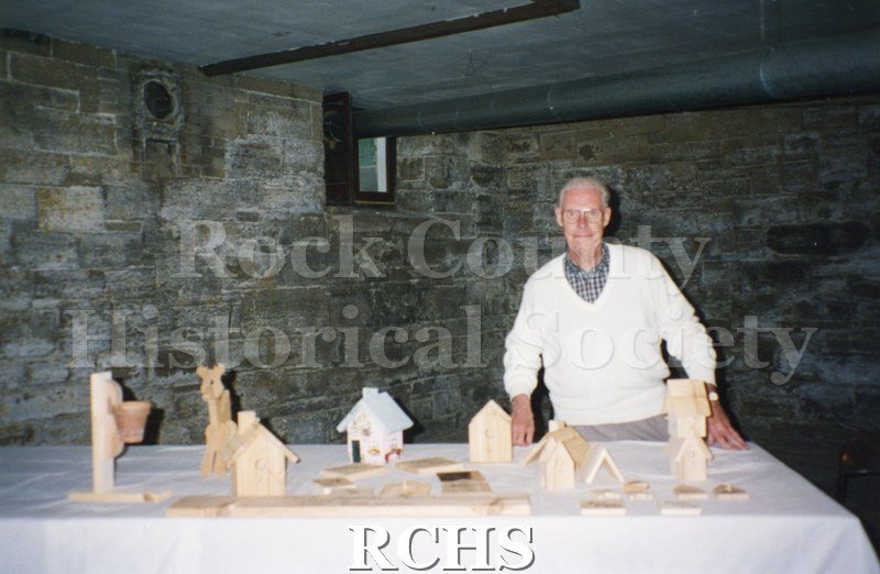 Color photo of Bob Wilson standing in front of table with wooden bird houses on it.