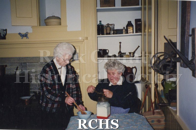 Color photo of Mabel Latzke and Arlene Ledger making butter. The one on the right is turning the churn and the other is stirring something in a bowl.