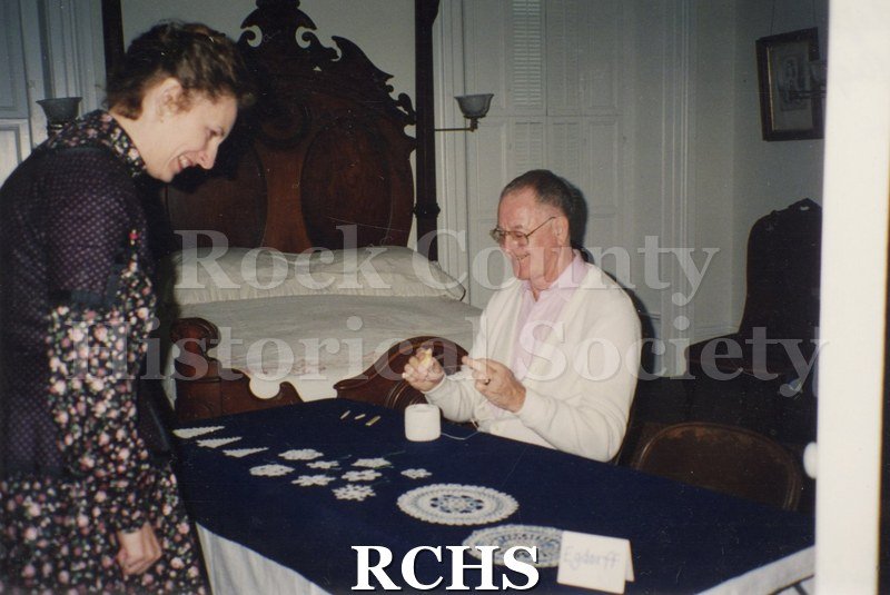Color photo of Joani Jarstad standing observing while Walter Egdorff is tatting.