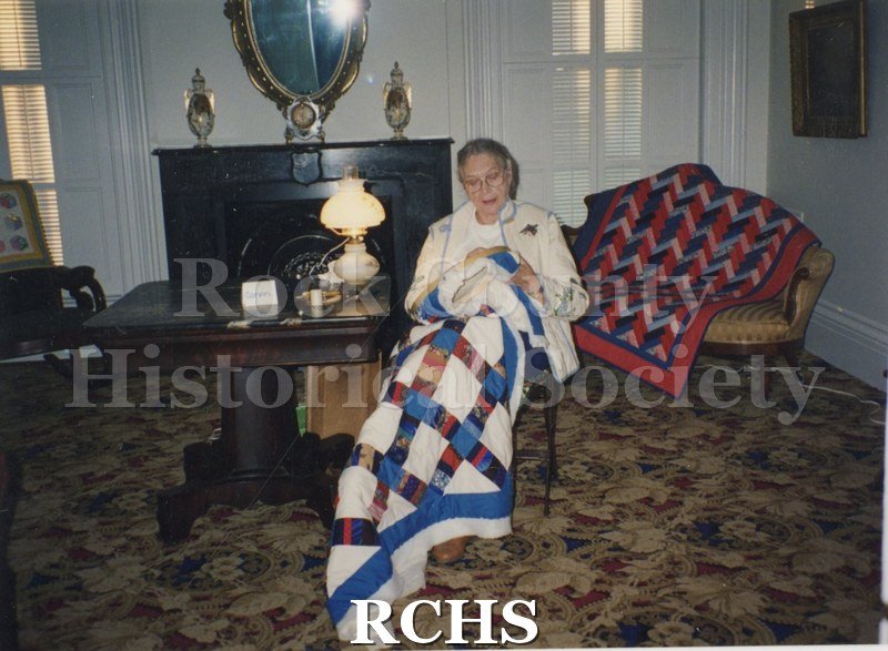 Photogrpah. Colored photograph of Ada Gavrin sitting in a chair while working on a quilt.