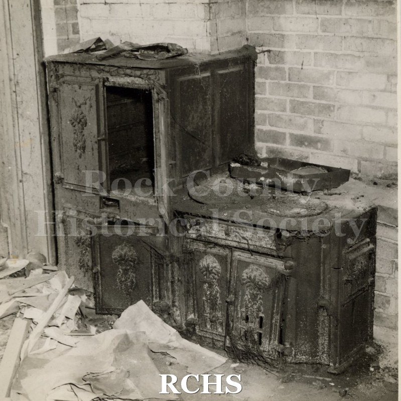 Black and white photograph of the Tallman House kitchen showing the brick fireplace with stove. There is debris on the floor.