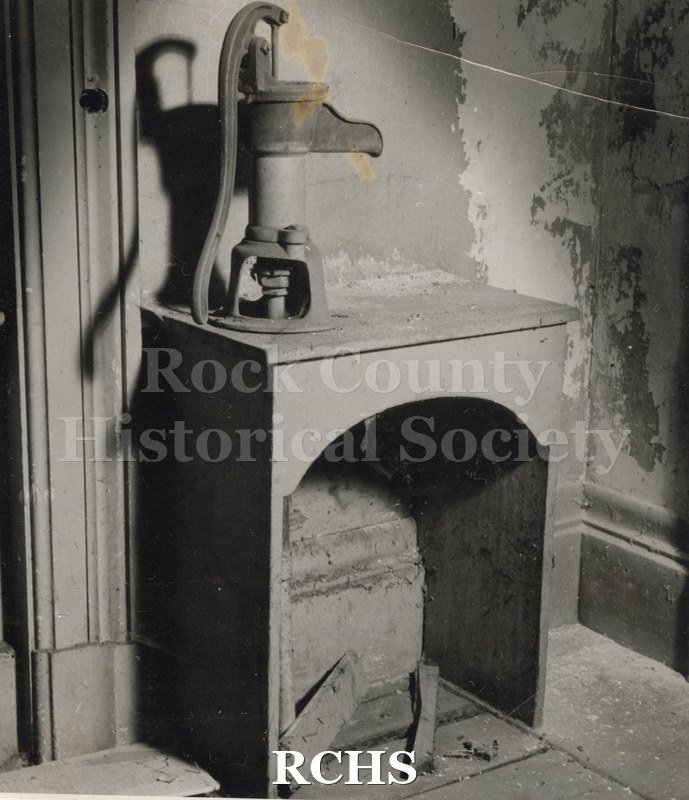 Black and white photograph of the Tallman House cistern pump in first floor kitchen. This is a hand pump on a stand. The wall is damaged.