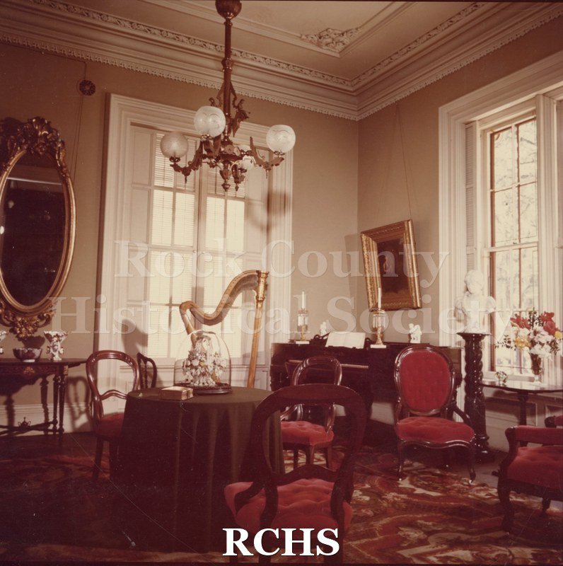 Color photo of the sitting room/parlor in the Tallman House. Shows the piano, harp, round table, and red upholstered chairs. c.1975.