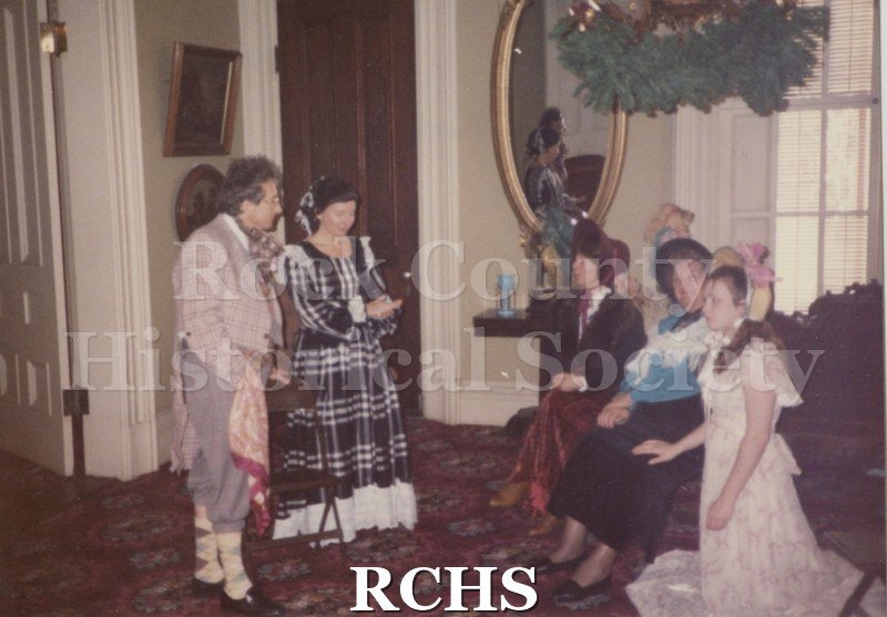 Historic image of Color photographof five unidentified people dressed in period clothing. Three are sitting on the settee and two are standing.