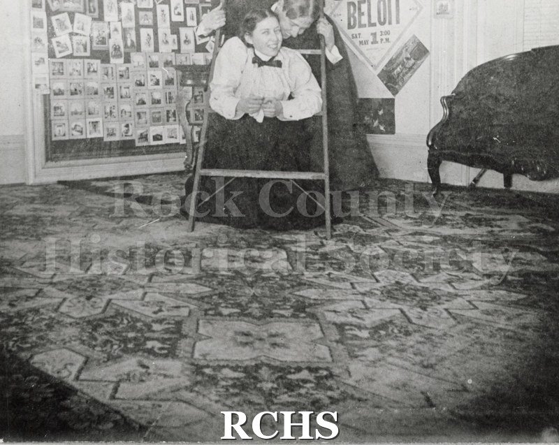 Historic image of Black ad white copy print of an unidentified man and woman. She is on her knees inside a ladder and the man is standing behind her.