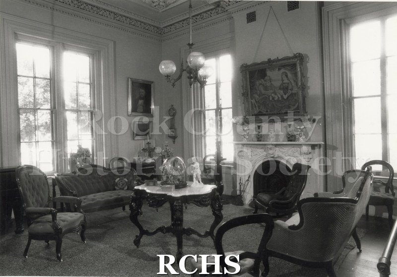 Black and white photograph of the sitting room of the Tallman House. Shows the fireplace with a painting above it; a marble topped table and chairs.
