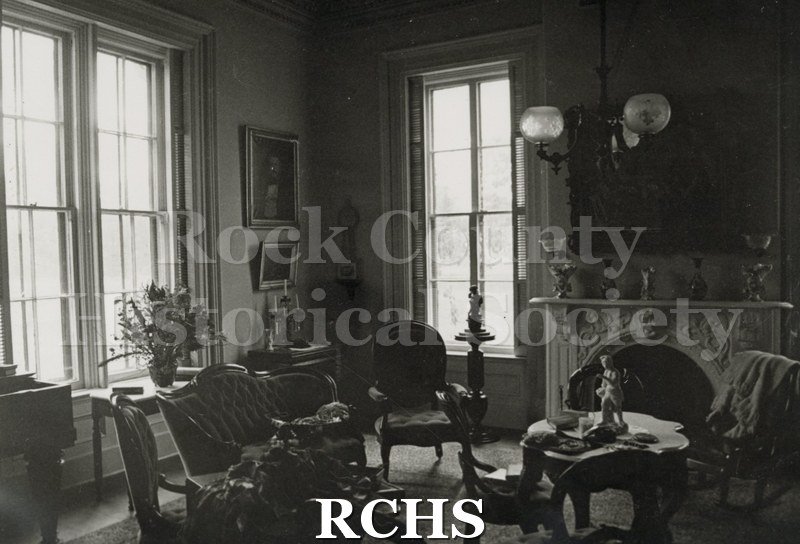 Black and white photograph of the sitting room of the Tallman House. Shows the fireplace with a painting above it; a marble topped table and chairs.
