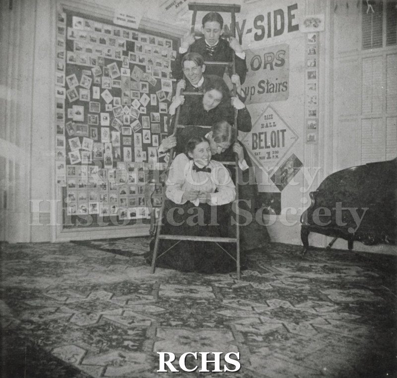 Historic image of Black and white copy print of five unidentified actors posed inbetween the steps of a ladder There are photographs on the wall and a sign: "Beloit.