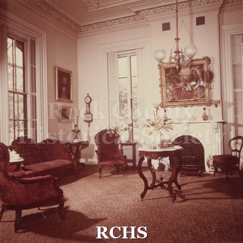 Color photo of the sitting room of the Tallman House. Shows the fireplace with a painting above it; a marble topped table and chairs.