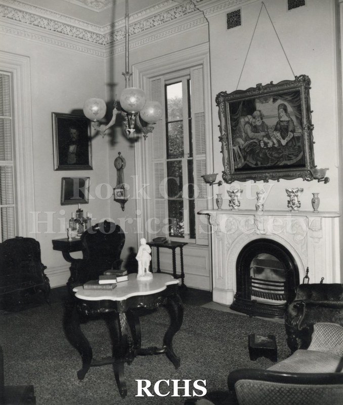 Black and white photograph of the sitting room of the Tallman House. Shows the fireplace with a painting above it; a marble topped table and chairs.