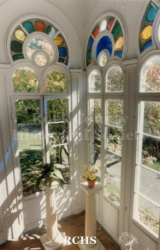 Color photo of the conservatory in the Tallman House showing three arched windows and the stained glass above them. There are two pedestals with a plant on each.
