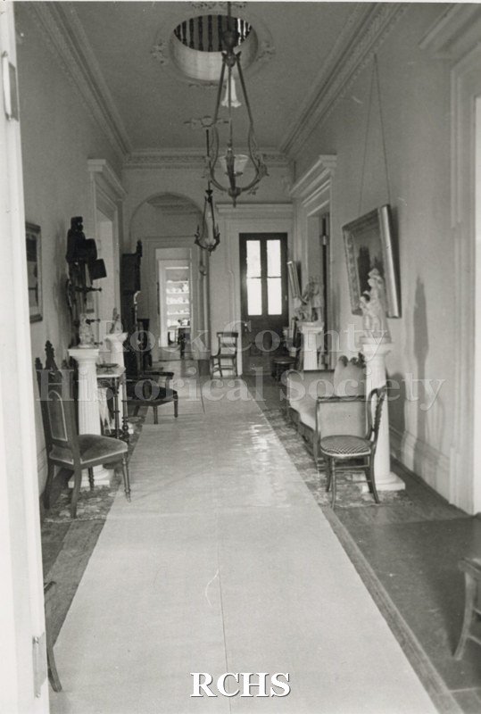 Black and white photograph of the front hall of the Tallman House showing pedestals with figures on them and chairs along the wall.