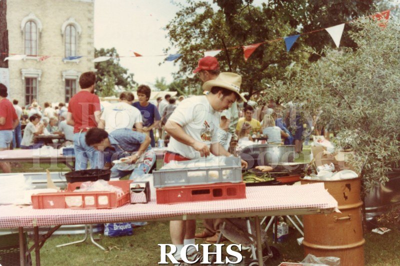 Color photo of a volunteer working on the food table. There is an overflowing trash barrel on the right.