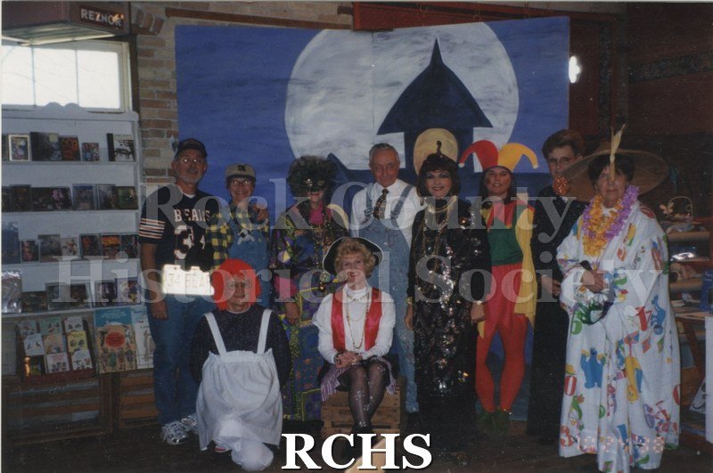 Color photo of Snap Apple Night. Shows several men and women in costumes posing in front of a backdrop with a moon and clock painted on it.